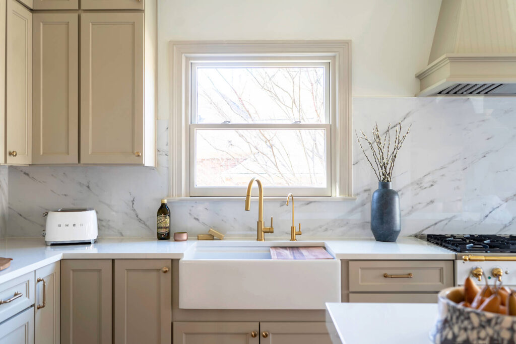 Timeless kitchen renovation with luxury design on a budget. View of the sink with tan painted cabinets, white countertops and a marble backsplash